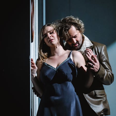 A performer wearing a navy-blue silk dress stands near a wall. Their eyes are closed, and their hands are being stroked from behind in a passionate embrace by another performer wearing a brown leather jacket and trousers and patterned beige shirt. His head almost rests gently on her shoulder.