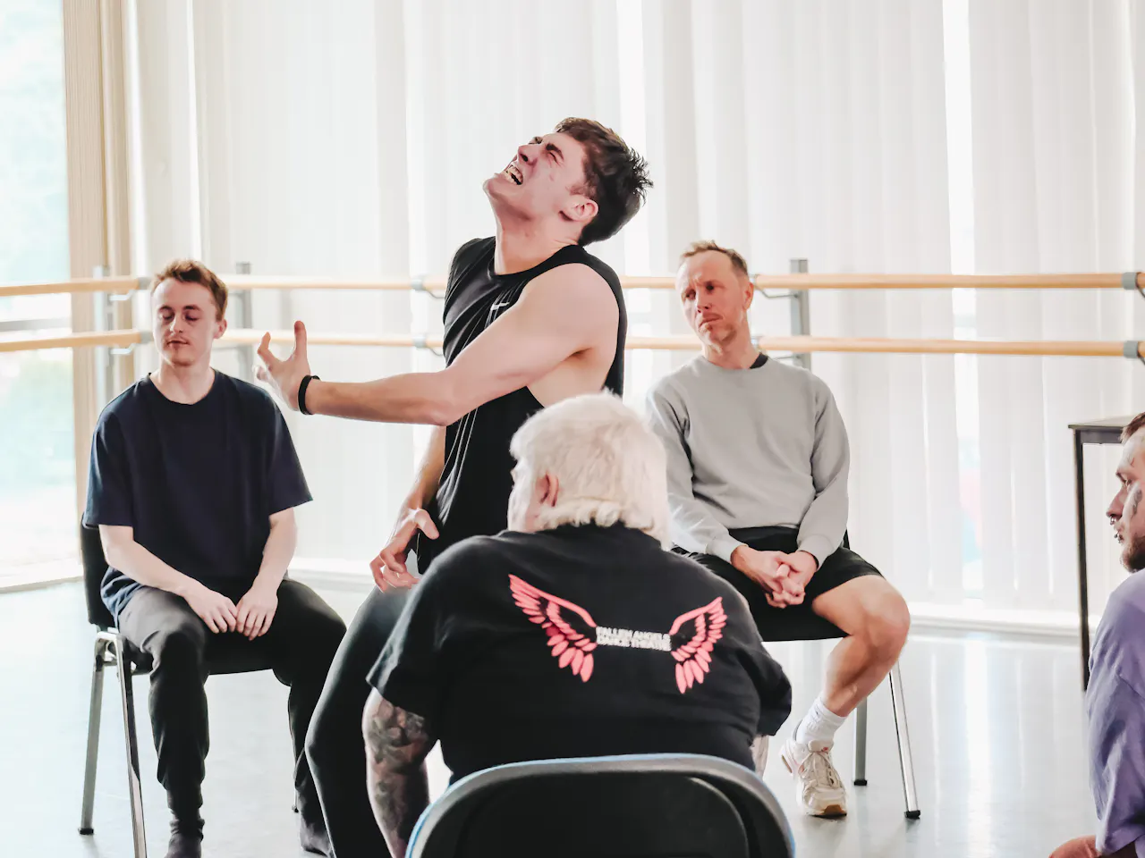 Four people sit in chairs in a brightly lit dance studio: two have their backs to the camera. In the centre, a dancer is performing – their face is contorted as though they are in pain, and they hold one arm out in front of themselves.