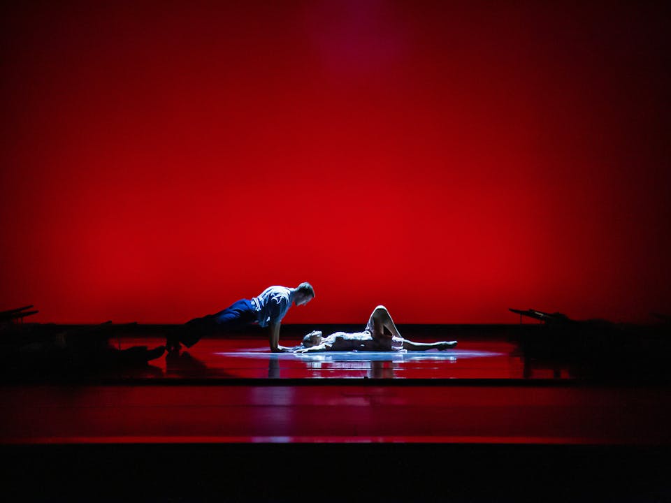 The stage is dark but set with a red backdrop with a spotlight focusing on two dancers. One dancer is lying on their back with one leg bent and their arms stretched upwards. Another dancer is staring into their eyes from above their head in a plank pose.