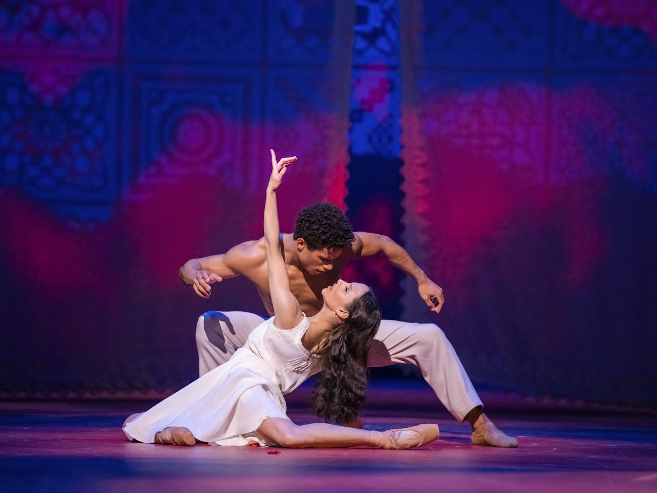 The ballet dancer Francesca Hayward wears a white dress and partially crouches on the floor with her left arm stretched upwards. She is leaning back with her long brown hair flowing downwards, almost to rest on the legs of the dancer Marcelino Sambé who is crouched down looking into her eyes. He is wearing only grey trousers. They are performing as Tita and Pedro in The Royal Ballet’s Like Water for Chocolate.