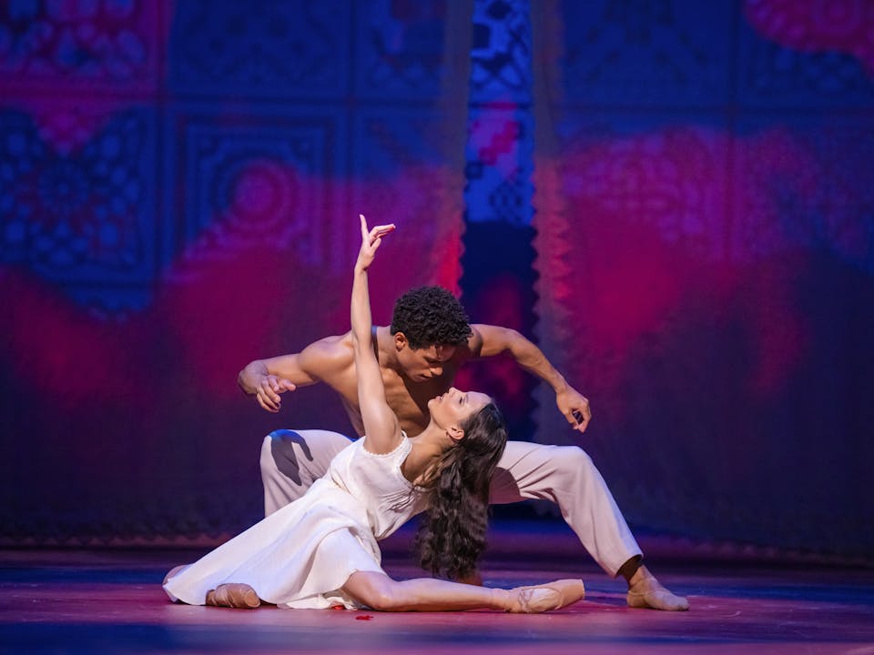 The ballet dancer Francesca Hayward wears a white dress and partially crouches on the floor with her left arm stretched upwards. She is leaning back with her long brown hair flowing downwards, almost to rest on the legs of the dancer Marcelino Sambé who is crouched down looking into her eyes. He is wearing only grey trousers. They are performing as Tita and Pedro in The Royal Ballet’s Like Water for Chocolate.