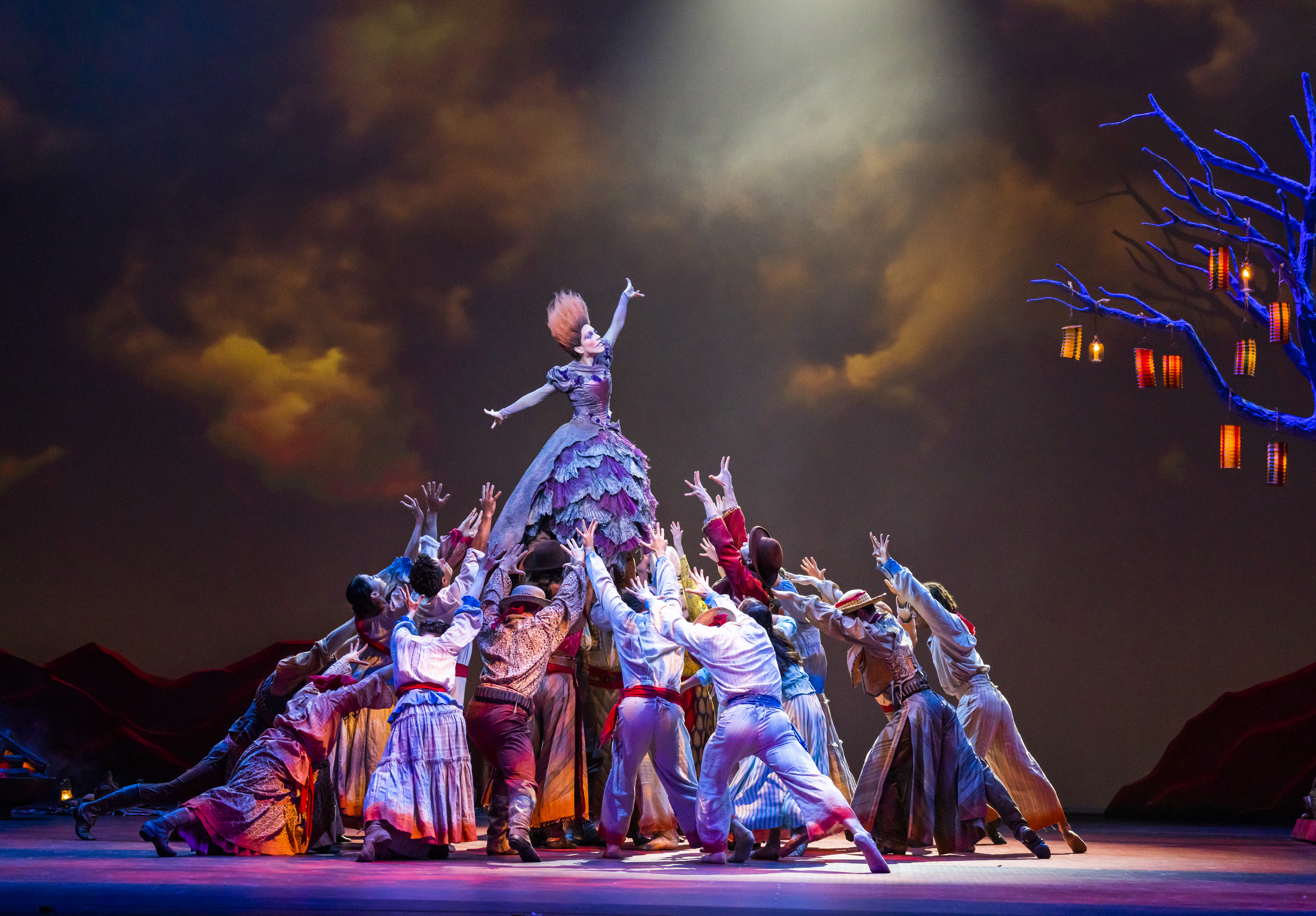 A dancer stands tall with their arms stretched outwards while being held up above the heads of dozens of performers. She wears a purple ballgown – the skirt of which is made up of layers of patchwork lace and her hair is orange and brushed upwards, so it sticks up. The other dancers are wearing colourful Hispanic peasant-style clothing. The stage is dark with clouds painted on the background but lit up by a skeletal tree lined with lanterns. 