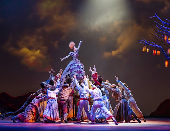 A dancer stands tall with their arms stretched outwards while being held up above the heads of dozens of performers. She wears a purple ballgown – the skirt of which is made up of layers of patchwork lace and her hair is orange and brushed upwards, so it sticks up. The other dancers are wearing colourful clothing. The stage is dark with clouds painted on the background but lit up by a skeletal tree lined with lanterns.