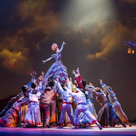 A dancer stands tall with their arms stretched outwards while being held up above the heads of dozens of performers. She wears a purple ballgown – the skirt of which is made up of layers of patchwork lace and her hair is orange and brushed upwards, so it sticks up. The other dancers are wearing colourful Hispanic peasant-style clothing. The stage is dark with clouds painted on the background but lit up by a skeletal tree lined with lanterns.