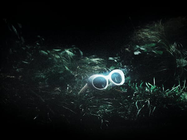 A pair of white 60s style framed sunglasses sit in amongst green grass. They represent the glasses worn by Kurt Cobain in The Royal Opera’s production of Last Days.