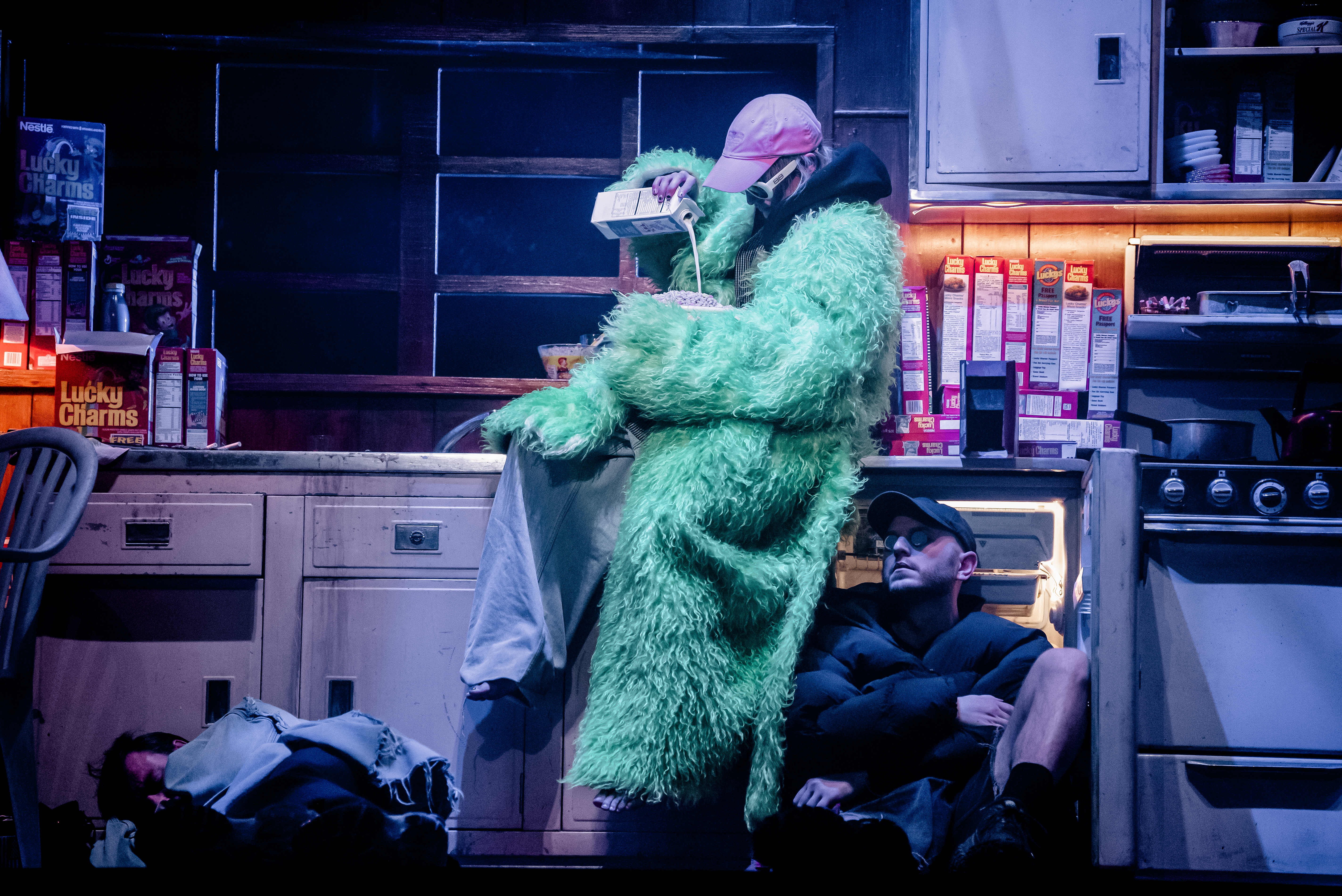 The stage is set to look like a neon-lit dirty kitchen filled with boxes of Lucky Charms cereal. A character wearing a green faux-fur coat, baggy taupe-coloured trousers, a pink baseball cap and white 60s style framed sunglasses sit on the counter pouring milk into a large bowl containing cereal. Below them sits another performer wearing a black puffer jacket, shorts, a black baseball cap and sunglasses. They are sitting against an open fridge. Another performer looks to be asleep on the floor with a blanket covering them over.