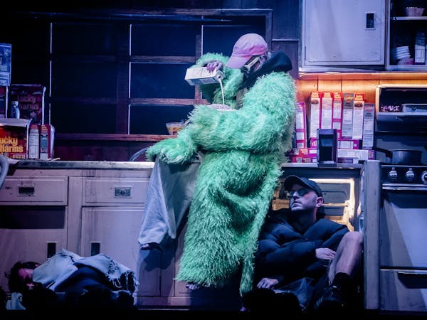 The stage is set to look like a neon-lit dirty kitchen filled with boxes of Lucky Charms cereal. A character wearing a green faux-fur coat, baggy taupe-coloured trousers, a pink baseball cap and white 60s style framed sunglasses sit on the counter pouring milk into a large bowl containing cereal. Below them sits another performer wearing a black puffer jacket, shorts, a black baseball cap and sunglasses. They are sitting against an open fridge. Another performer looks to be asleep on the floor with a blanket covering them over.