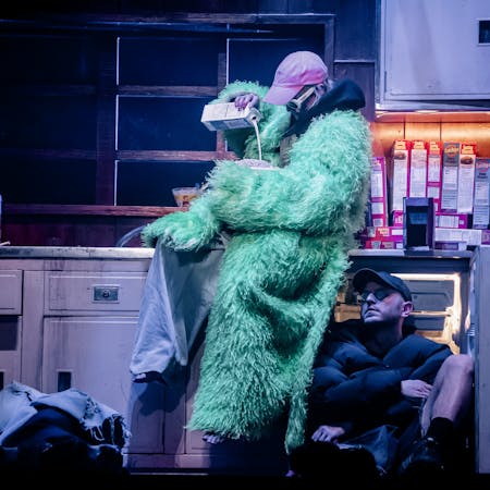 The stage is set to look like a neon-lit dirty kitchen filled with boxes of Lucky Charms cereal. A character wearing a green faux-fur coat, baggy taupe-coloured trousers, a pink baseball cap and white 60s style framed sunglasses sit on the counter pouring milk into a large bowl containing cereal. Below them sits another performer wearing a black puffer jacket, shorts, a black baseball cap and sunglasses. They are sitting against an open fridge. Another performer looks to be asleep on the floor with a blanket covering them over.
