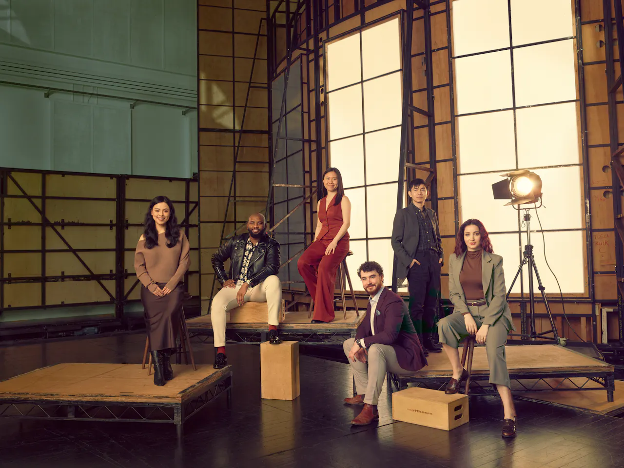 Six people who are members of the Jette Parker Artists sit or stand on raised wooden platforms of varying heights in front of a large lightbox made to look like a window in a photography studio.