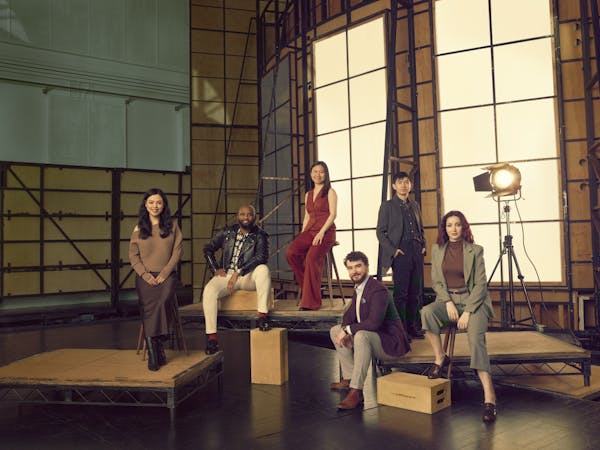 Six people who are members of the Jette Parker Artists sit or stand on raised wooden platforms of varying heights in front of a large lightbox made to look like a window in a photography studio.