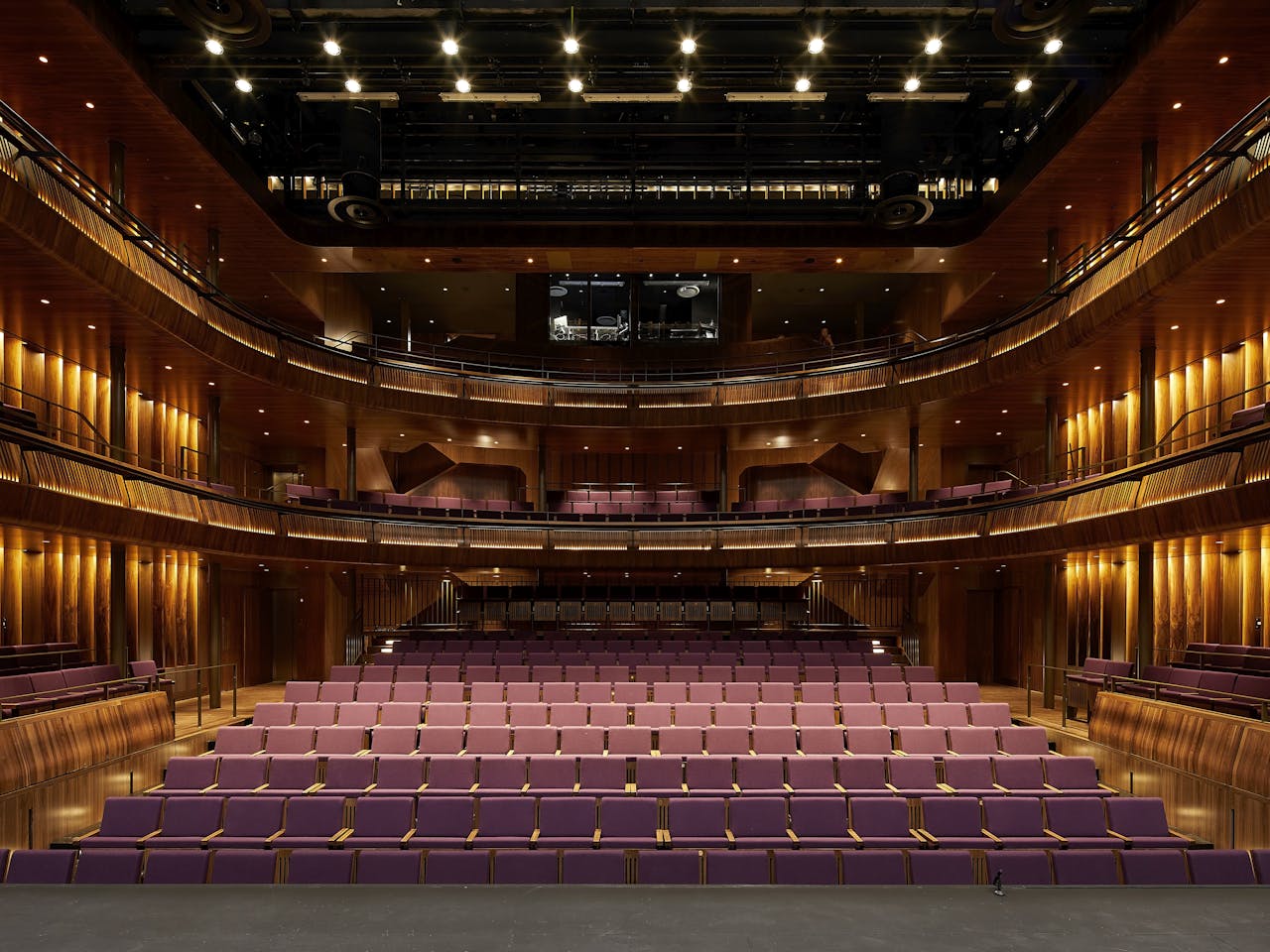 A view across the Linbury Stage auditorium of The Royal Opera House from the stage. It is empty and well-lit showing the golden-coloured wooden panelling and purple seating of the auditorium. The light and sound control box that sits at the top of the auditorium is visible as a brightly lit room.