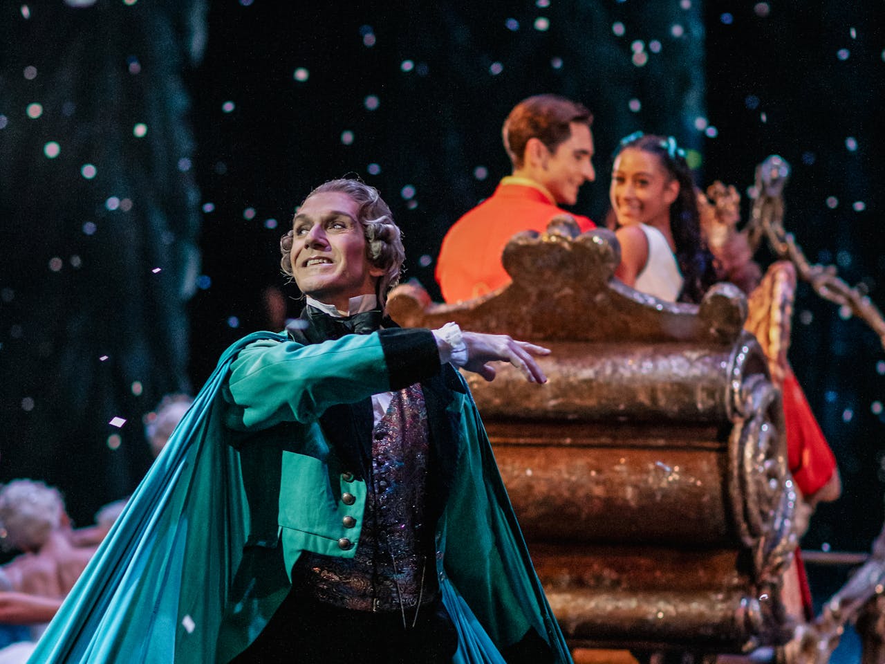 The ballet dancer Gary Avis performs as Herr Drosselmeyer in The Royal Ballet’s Nutcracker – he wears a grey powder wig, a white frilled shirt, a green bow tie, sparkly colourful waistcoat and a teal-coloured clothing cloak. Behind him is a gold carriage carrying two people who are looking out towards him. They are dancers Giacomo Rovero and Marianna Tsembenhoi who are performing as Hans Peter and Clara.