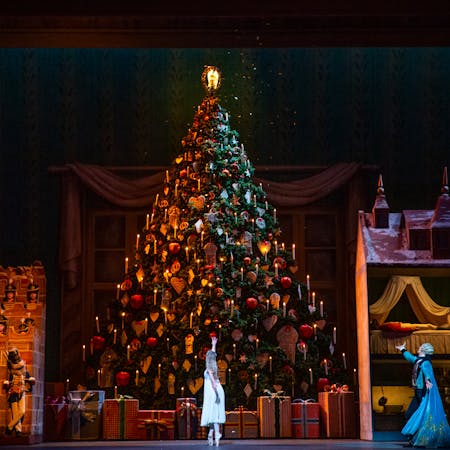 An enormous, decorated Christmas tree stands in the centre of the stage. It is lined with large beautifully wrapped presents bound with ribbon. To the left of the tree stands a large toy-like castle with several performers dressed as soldiers standing inside. To the other side sits a toy-like dollhouse with a solider lying on the bed in one room. In front, the ballet dancer Gary Avis performing as Herr Drosslemeyer holds his arm out as another dancer wearing a white dress performing as Clara stands en pointe with her arm reaching upwards.