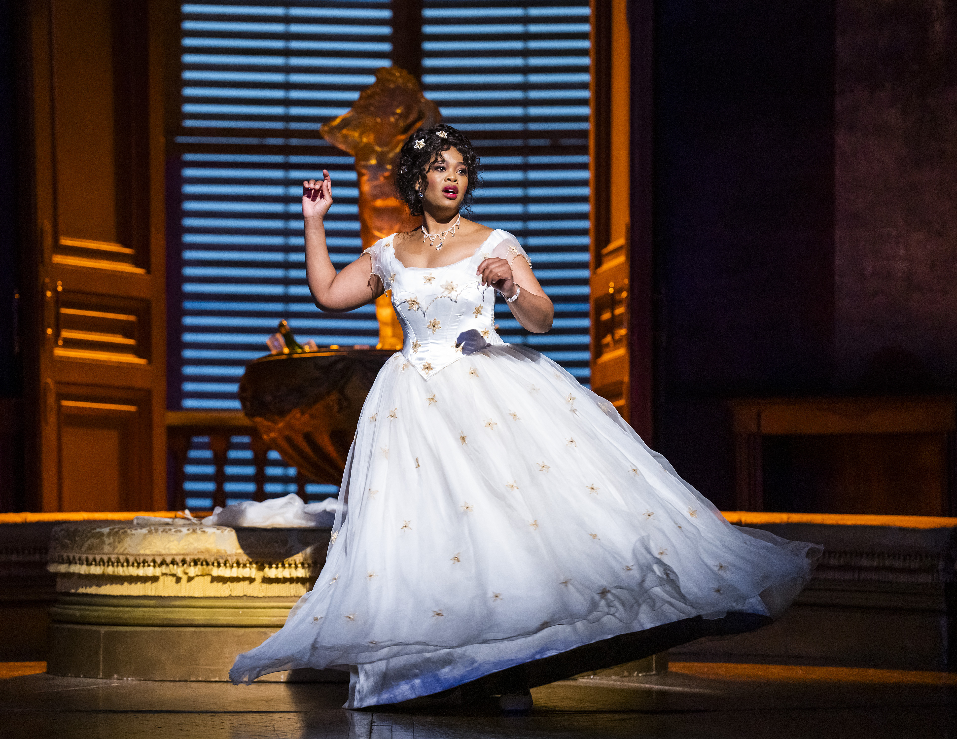 The operatic soprano Pretty Yende wears a white ballgown embroidered with yellow flowers as she stands in a sumptuous looking room with a large, shuttered window behind her. She is performing as Violetta Valéry in Thye Royal Opera’s production of La traviata.  