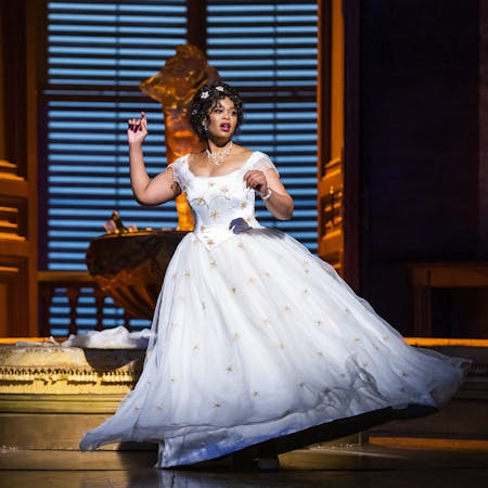 The operatic soprano Pretty Yende wears a white ballgown embroidered with yellow flowers as she stands in a sumptuous looking room with a large, shuttered window behind her. She is performing as Violetta Valéry in Thye Royal Opera’s production of La traviata.