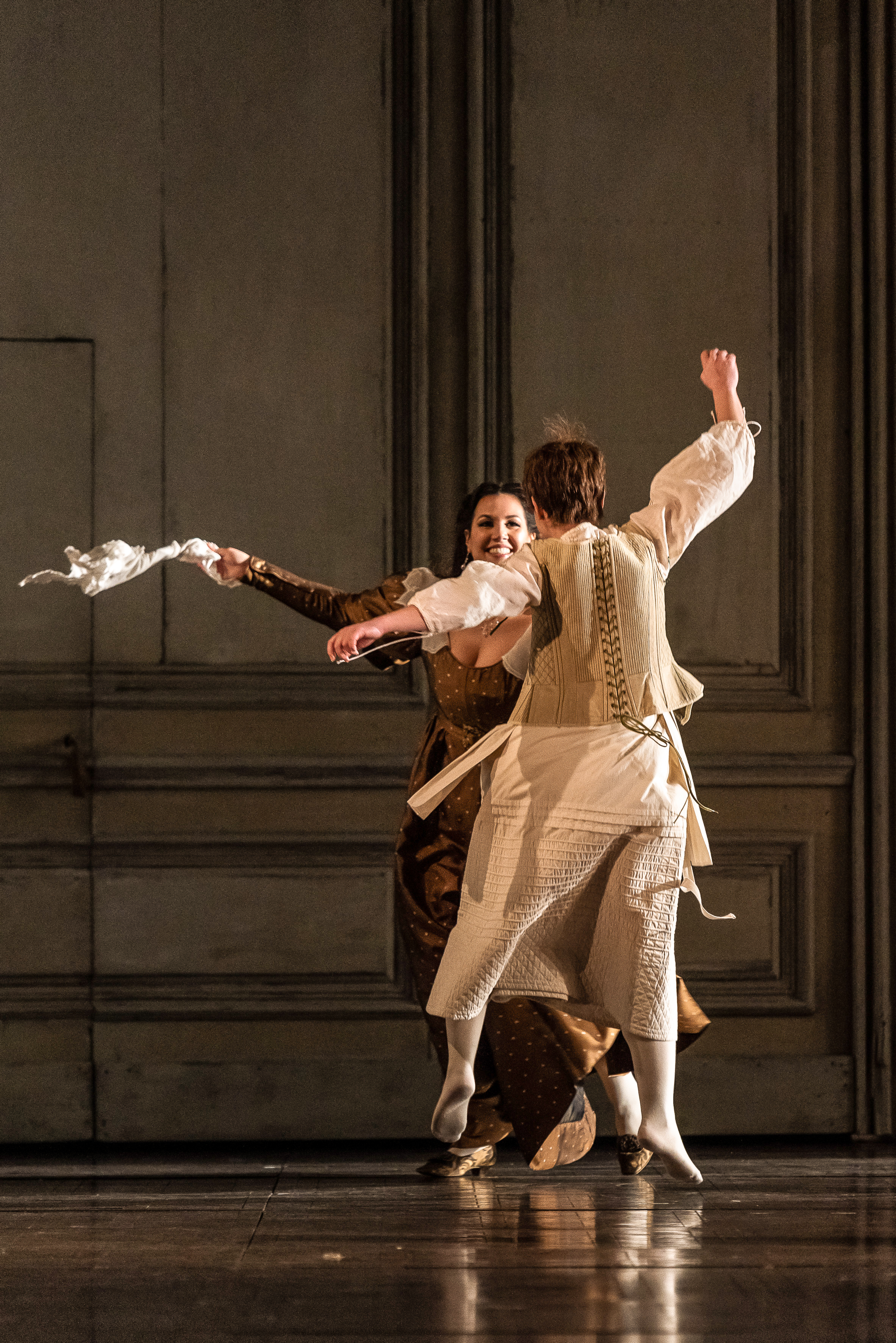 Two performers dance together in The Royal Opera’s production of The Marriage of Figaro. One is facing the audience, and she is wearing a long brown dress. A white cloth she is holding is flying in the air as she moves her arms. The other performer wears a white shirt, underclothes and white tights. They have short blonde hair.  