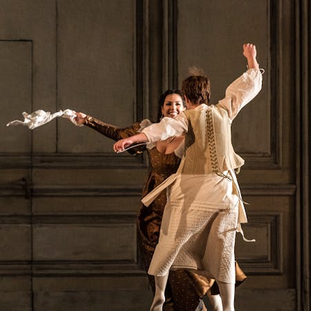 Two performers dance together in The Royal Opera’s production of The Marriage of Figaro. One is facing the audience, and she is wearing a long brown dress. A white cloth she is holding is flying in the air as she moves her arms. The other performer wears a white shirt, underclothes and white tights. They have short blonde hair.