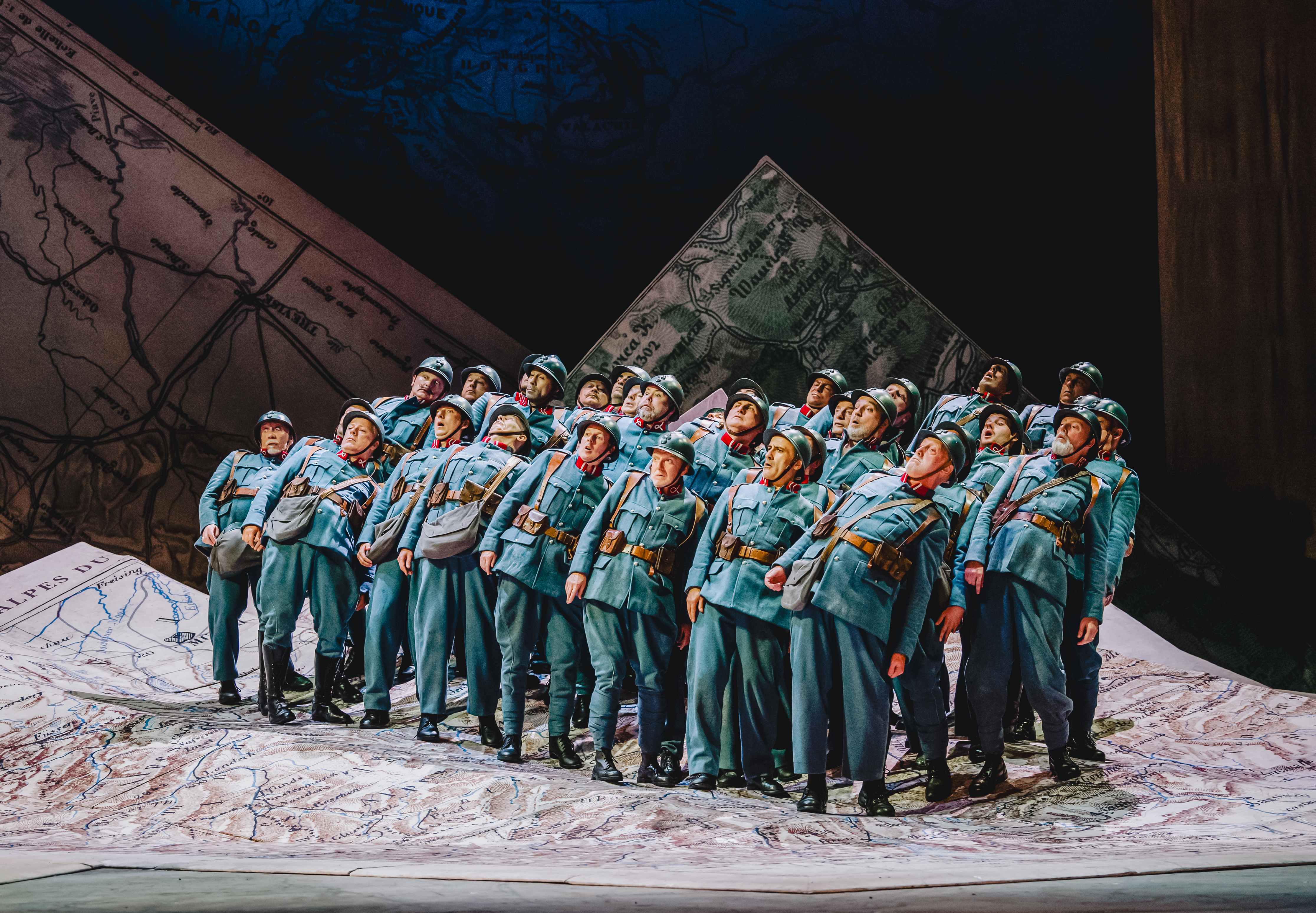Members of The Royal Opera Chorus perform in The Royal Opera’s production of La fille du régiment. They are wearing blue army uniforms and helmets and standing together on a large, unfolded map. They all lean backwards.   