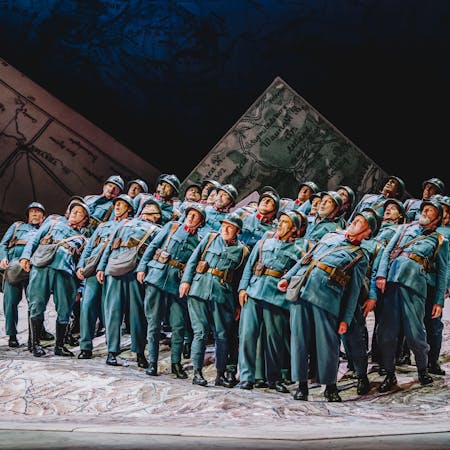Members of The Royal Opera Chorus perform in The Royal Opera’s production of La fille du régiment. They are wearing blue army uniforms and helmets and standing together on a large, unfolded map. They all lean backwards.