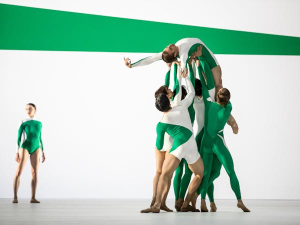 A ballet dancer wearing a green and white striped leotard is being held up by their shoulders, back and legs by five other ballet dancers, dressed similarly. They are on their back facing upwards with one arm stretched out above their head. In the background, another dancer wearing a similarly patterned leotard stands tall looking over. The stage is set with green and white stripes that match the dancers' leotards.
