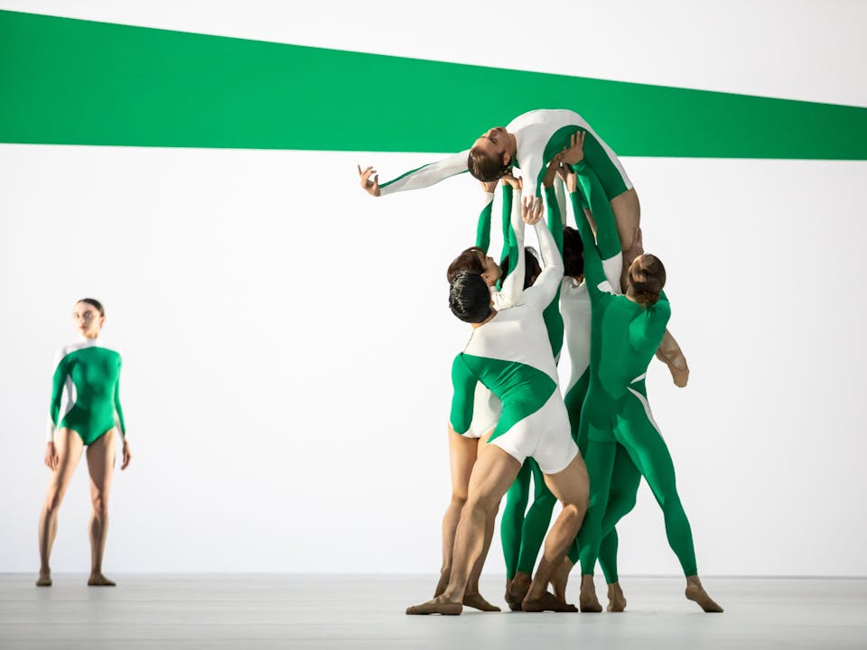 A ballet dancer wearing a green and white striped leotard is being held up by their shoulders, back and legs by five other ballet dancers, dressed similarly. They are on their back facing upwards with one arm stretched out above their head. In the background, another dancer wearing a similarly patterned leotard stands tall looking over. The stage is set with green and white stripes that match the dancers' leotards.