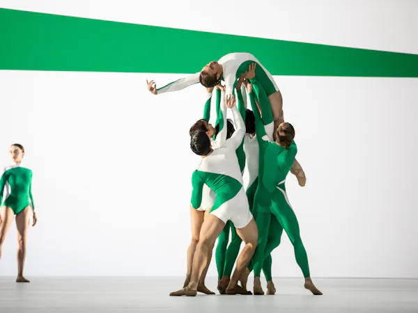 A ballet dancer wearing a green and white striped leotard is being held up by their shoulders, back and legs by five other ballet dancers, dressed similarly. They are on their back facing upwards with one arm stretched out above their head. In the background, another dancer wearing a similarly patterned leotard stands tall looking over. The stage is set with green and white stripes that match the dancers' leotards.