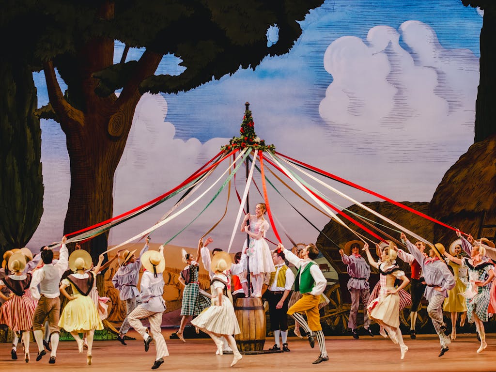 Dozens of dancers dressed in colourful striped clothing and bonnets dance around a maypole, holding on to the red, green and white ribbons that flow down from the top of the pole. A ballet dancer wearing a pink and white corseted dress, white tights and ballet slippers stands on a wooden barrel, holding on to the pole. The stage background is set with painted rural scenery of trees and haystacks.