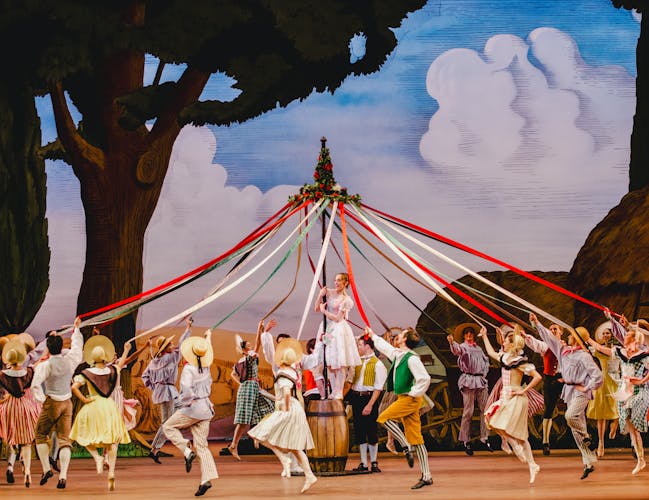 Dozens of dancers dressed in colourful striped clothing and bonnets dance around a maypole, holding on to the red, green and white ribbons that flow down from the top of the pole. A ballet dancer wearing a pink and white corseted dress, white tights and ballet slippers stands on a wooden barrel, holding on to the pole. The stage background is set with painted rural scenery of trees and haystacks.