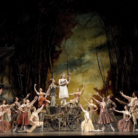 The ballet dancer Marianela Nuñez, performing as Giselle, stands on a wooden carriage draped with grapes. She is wearing a brown corset over a cream dress and a flower headdress and holds up twigs and flowers. A dancer wearing a brown jacket, boots and belt puts one arm around her. Dozens of dancers stand or kneel around the carriage holding one arm up towards her. The scenery on the stage is set to look like a rural-style brushwork painting with cottages and trees.