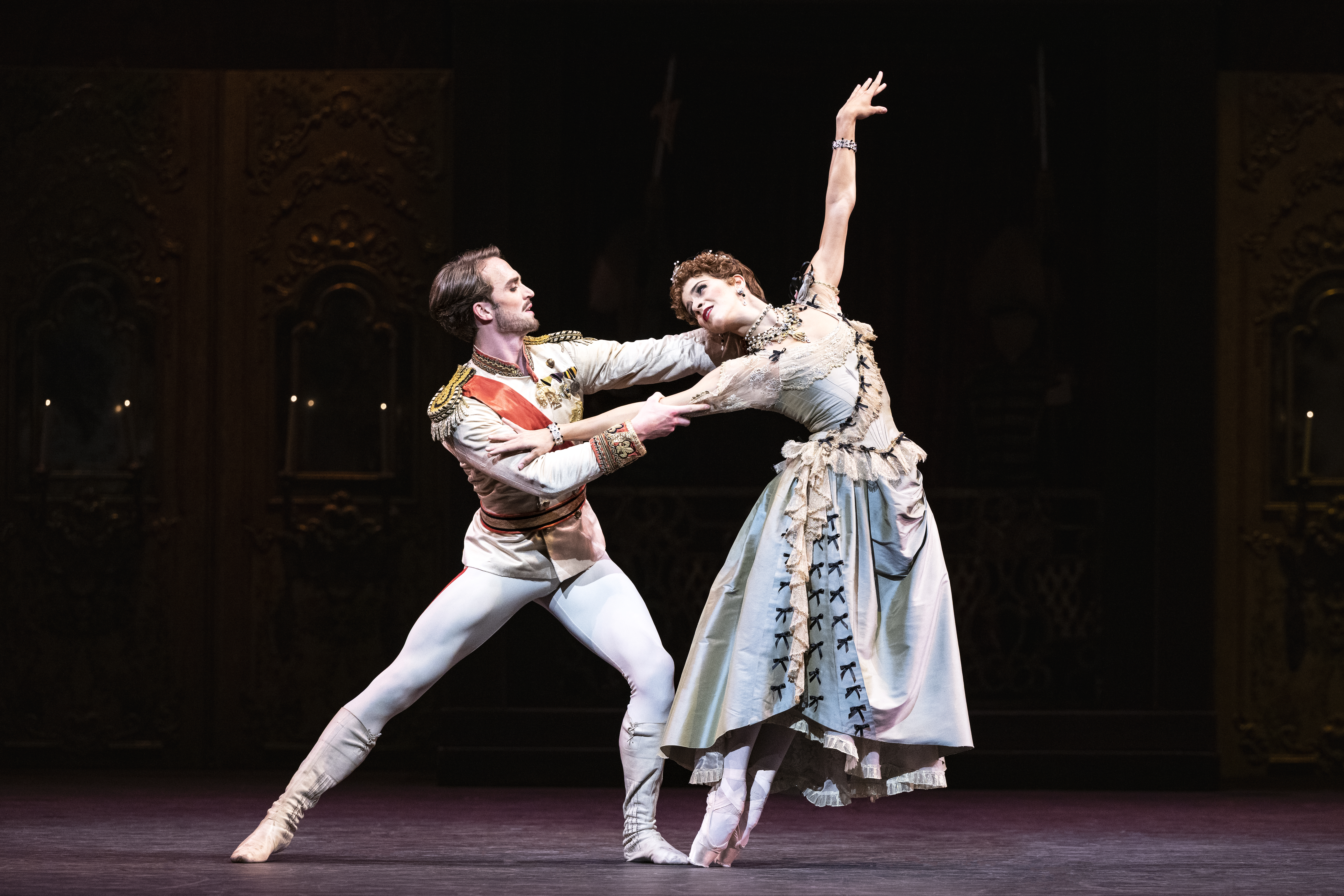 The ballet dancer Matthew Ball is performing as Crown Prince Rudolf in The Royal Ballet’s production of Kenneth MacMillan’s Mayerling. He wears a white soldier's uniform with a red sash and there are medals on his chest. He is holding a pose in a lunging position with his arms stretched out to the shoulders of Mayara Magri who is performing as Countess Marie Larisch. One arm is stretched outwards, and the other is raised above her head, and she is en pointe. She is wearing a light blue satin and lace dress with a corset. The dress has black bows embroidered across from her right shoulder to her left leg.  