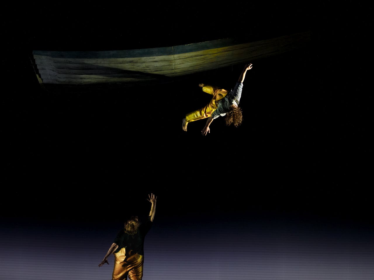 The tenor Allan Clayton stands with his back to the audience on stage performing the role of Peter Grimes in The Royal Opera’s Peter Grimes. He is wearing yellow fishing overalls and holds one arm upwards and looks up. Above, a performer wearing the same yellow fishing overalls appears to dangle by one foot in the air, their long brown curly hair draping over their face as they are suspended.