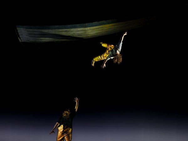 The tenor Allan Clayton stands with his back to the audience on stage performing the role of Peter Grimes in The Royal Opera’s Peter Grimes. He is wearing yellow fishing overalls and holds one arm upwards and looks up. Above, a performer wearing the same yellow fishing overalls appears to dangle by one foot in the air, their long brown curly hair draping over their face as they are suspended.