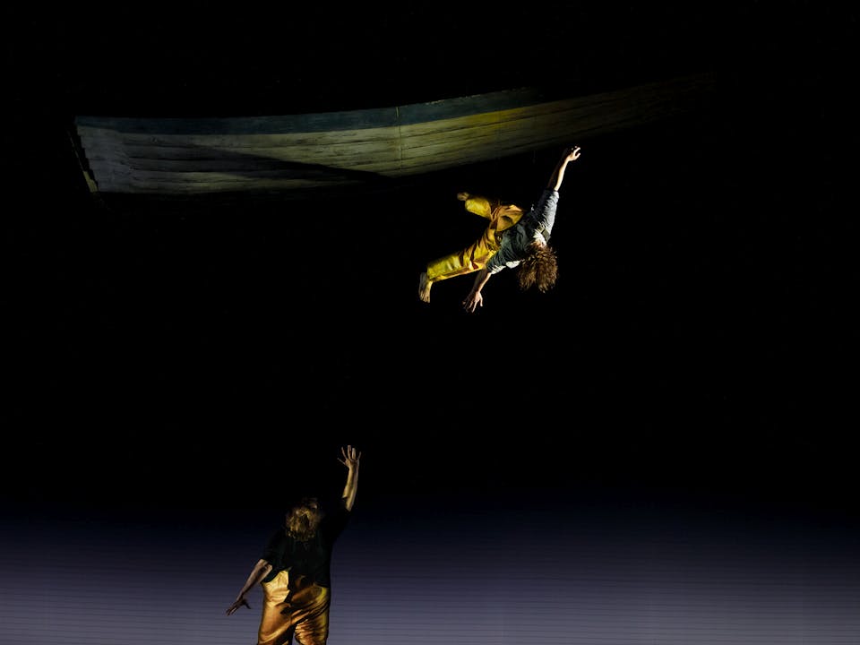 The tenor Allan Clayton stands with his back to the audience on stage performing the role of Peter Grimes in The Royal Opera’s Peter Grimes. He is wearing yellow fishing overalls and holds one arm upwards and looks up. Above, a performer wearing the same yellow fishing overalls appears to dangle by one foot in the air, their long brown curly hair draping over their face as they are suspended.