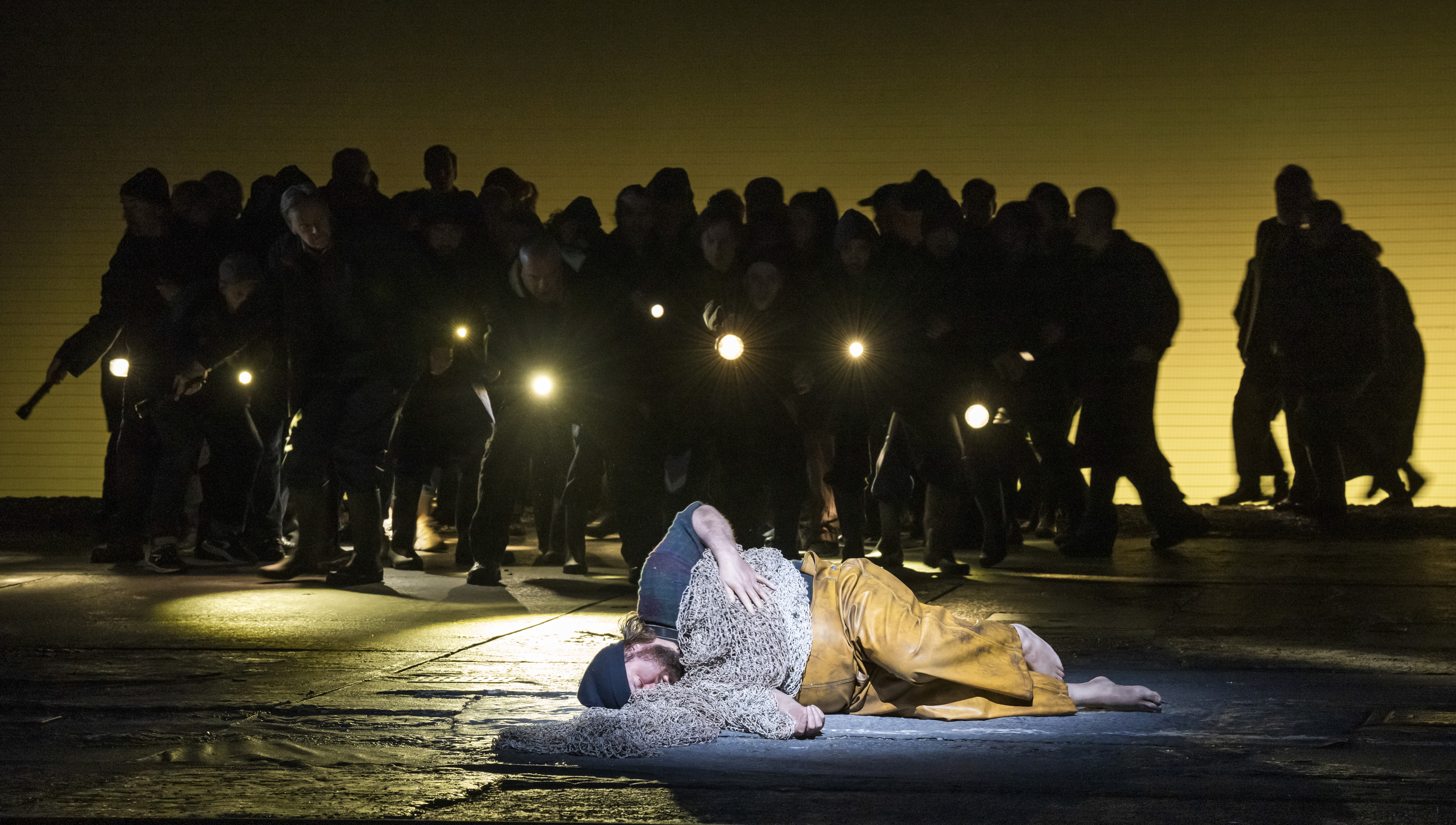 The tenor Allan Clayton is performing as Peter Grimes in The Royal Opera’s Peter Grimes. He is barefoot, lying on the stage dressed in dirty yellow fishing overalls, a dark blue plaid shirt and a navy beanie hat, as though he is asleep. Fishing nets are draped over his body like a blanket. Behind him a chorus of performers stands in darkness holding torches.  