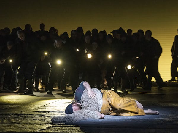 The tenor Allan Clayton is performing as Peter Grimes in The Royal Opera’s Peter Grimes. He is barefoot, lying on the stage dressed in dirty yellow fishing overalls, a dark blue plaid shirt and a navy beanie hat, as though he is asleep. Fishing nets are draped over his body like a blanket. Behind him a chorus of performers stands in darkness holding torches.