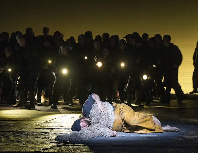 The tenor Allan Clayton is performing as Peter Grimes in The Royal Opera’s Peter Grimes. He is barefoot, lying on the stage dressed in dirty yellow fishing overalls, a dark blue plaid shirt and a navy beanie hat, as though he is asleep. Fishing nets are draped over his body like a blanket. Behind him a chorus of performers stands in darkness holding torches.