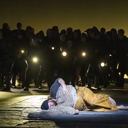 The tenor Allan Clayton is performing as Peter Grimes in The Royal Opera’s Peter Grimes. He is barefoot, lying on the stage dressed in dirty yellow fishing overalls, a dark blue plaid shirt and a navy beanie hat, as though he is asleep. Fishing nets are draped over his body like a blanket. Behind him a chorus of performers stands in darkness holding torches.