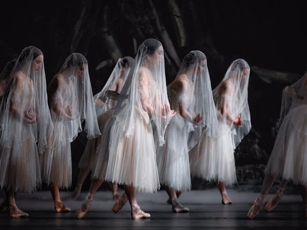 Nine ballet dancers stand on stage. They are all wearing the same white lace dress, pink pointe slippers, and a white translucent veil that is draped over their heads giving them ghostly appearances. They also have small translucent jeweled wings on their backs. They all hold their arms in front of them, their hands crossing over their wrists. They are Artists of The Royal Ballet performing in Giselle.