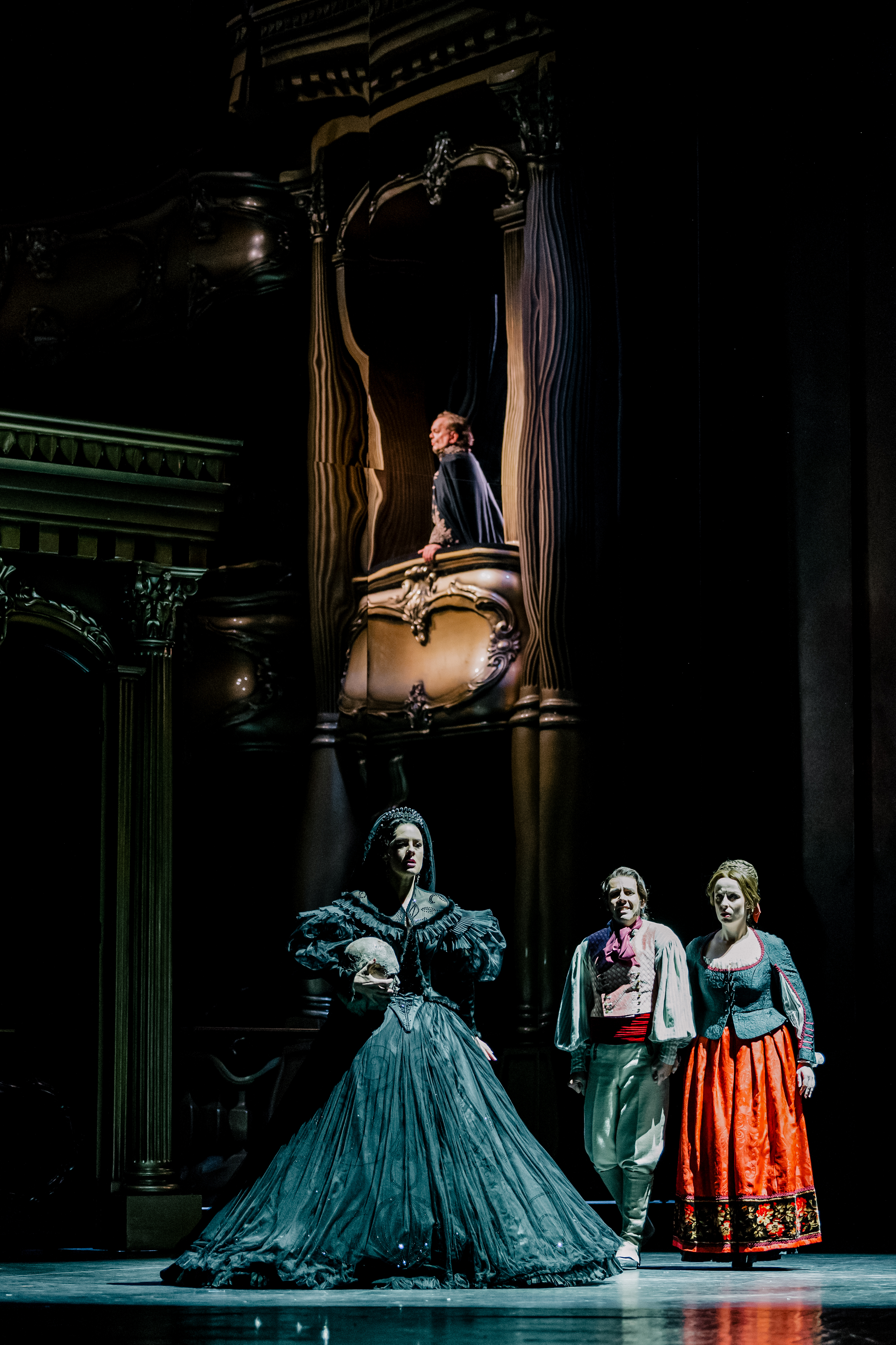 The soprano Malin Byström performs as Hélène in The Royal Opera’s production of The Sicilian Vespers. She stands in the centre of the stage wearing a large black ballgown and veil, holding a replica human skull in her right arm. Above her a performer wearing a blue cloak looks out across the stage from an opera box. Two performers wearing bright clothing look out to the audience.  