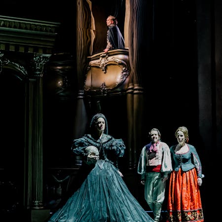 The soprano Malin Byström performs as Hélène in The Royal Opera’s production of The Sicilian Vespers. She stands in the centre of the stage wearing a large black ballgown and veil, holding a replica human skull in her right arm. Above her a performer wearing a blue cloak looks out across the stage from an opera box. Two performers wearing bright clothing look out to the audience.