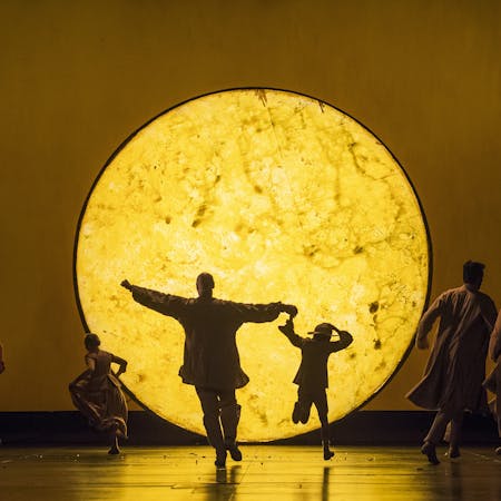 A large round yellow light replicating the moon is set in the centre of the stage. A person and a small child with their backs to the audience appear to run towards the centre of the moon.