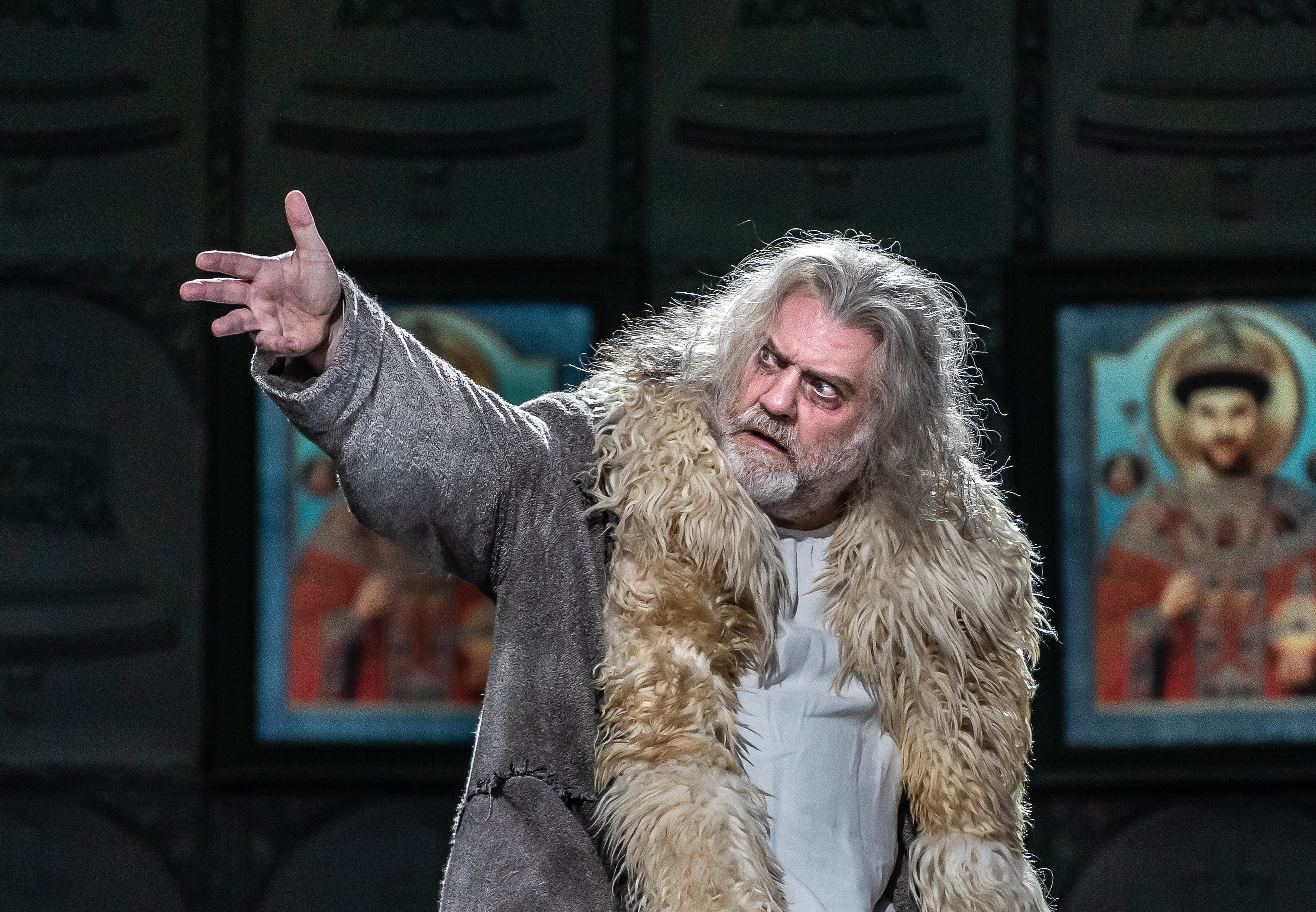 Bryn Terfel performs as Boris Godunov in The Royal Opera’s production of Boris Godunov. He has long grey hair and wears a coat lined with faux fur. He holds up his right hand and his eyes are wide conveying strong emotions. Several colourful iconographic paintings are visible from behind him.  