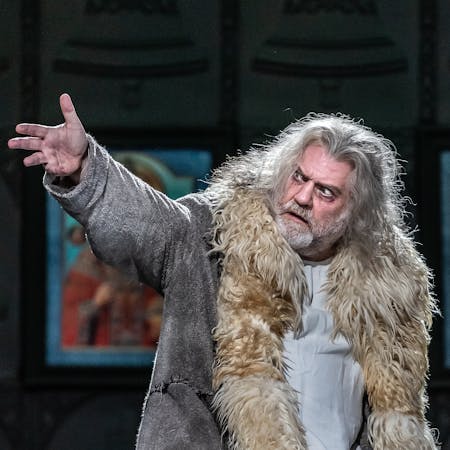Bryn Terfel performs as Boris Godunov in The Royal Opera’s production of Boris Godunov. He has long grey hair and wears a coat lined with faux fur. He holds up his right hand and his eyes are wide conveying strong emotions. Several colourful iconographic paintings are visible from behind him.