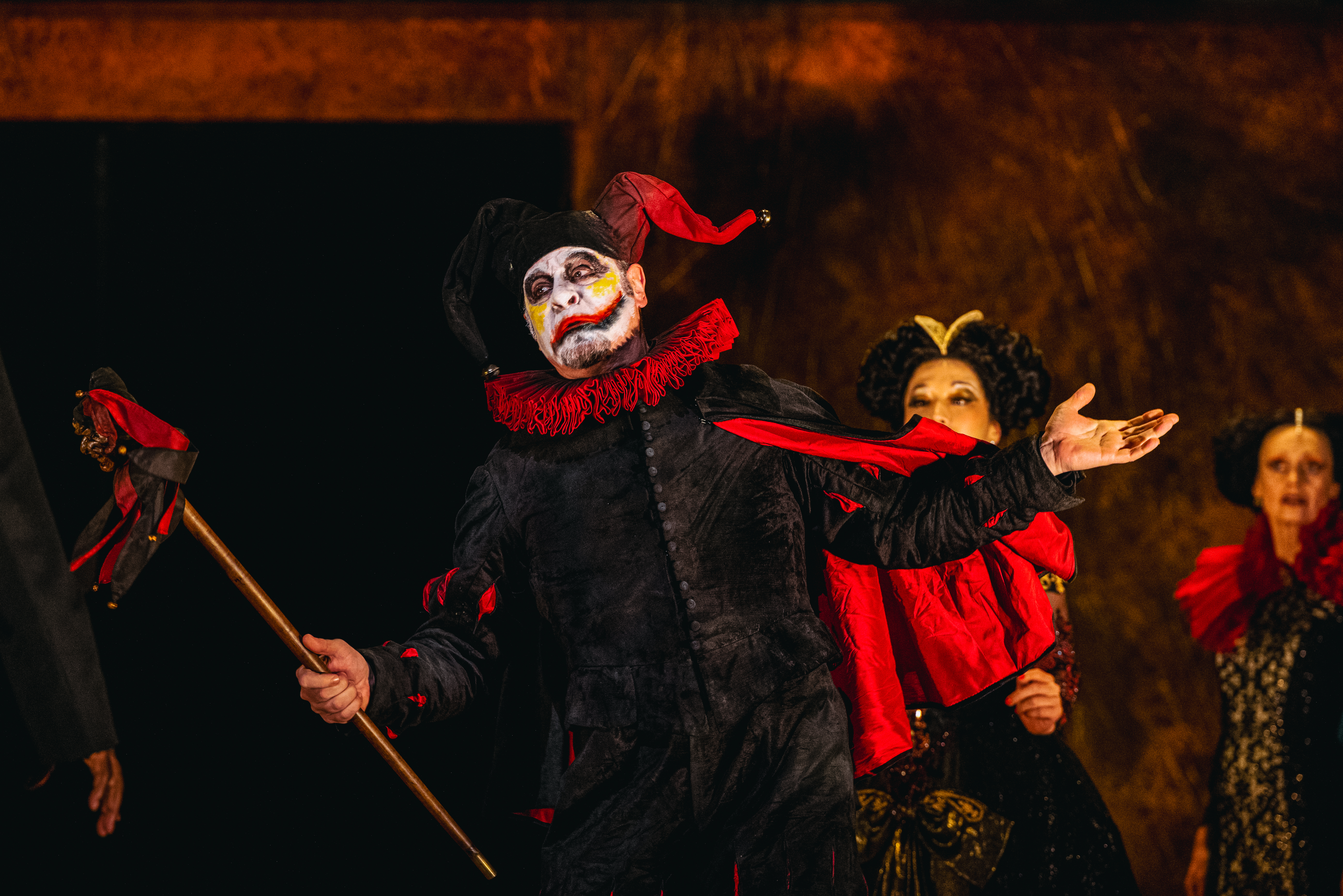 Carlos Álvarez performs as Rigoletto in The Royal Opera’s production of Rigoletto. A jester wearing a black suit, red ruffle and black and red jester's hat and he holds up a black and red marotte. His face is painted white with yellow marks on his cheeks and a black and red lip painted across his face. There are several performers standing behind him dressed in large gowns and wigs.  