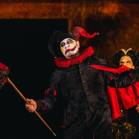 Carlos Álvarez performs as Rigoletto in The Royal Opera’s production of Rigoletto. A jester wearing a black suit, red ruffle and black and red jester's hat and he holds up a black and red marotte. His face is painted white with yellow marks on his cheeks and a black and red lip painted across his face. There are several performers standing behind him dressed in large gowns and wigs.