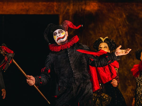 Carlos Álvarez performs as Rigoletto in The Royal Opera’s production of Rigoletto. A jester wearing a black suit, red ruffle and black and red jester's hat and he holds up a black and red marotte. His face is painted white with yellow marks on his cheeks and a black and red lip painted across his face. There are several performers standing behind him dressed in large gowns and wigs.