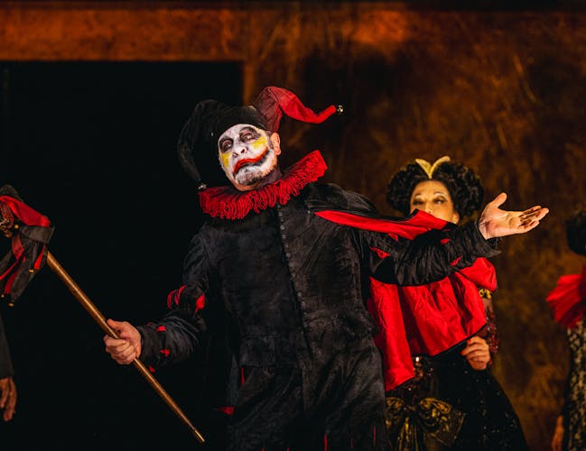 Carlos Álvarez performs as Rigoletto in The Royal Opera’s production of Rigoletto. A jester wearing a black suit, red ruffle and black and red jester's hat and he holds up a black and red marotte. His face is painted white with yellow marks on his cheeks and a black and red lip painted across his face. There are several performers standing behind him dressed in large gowns and wigs.