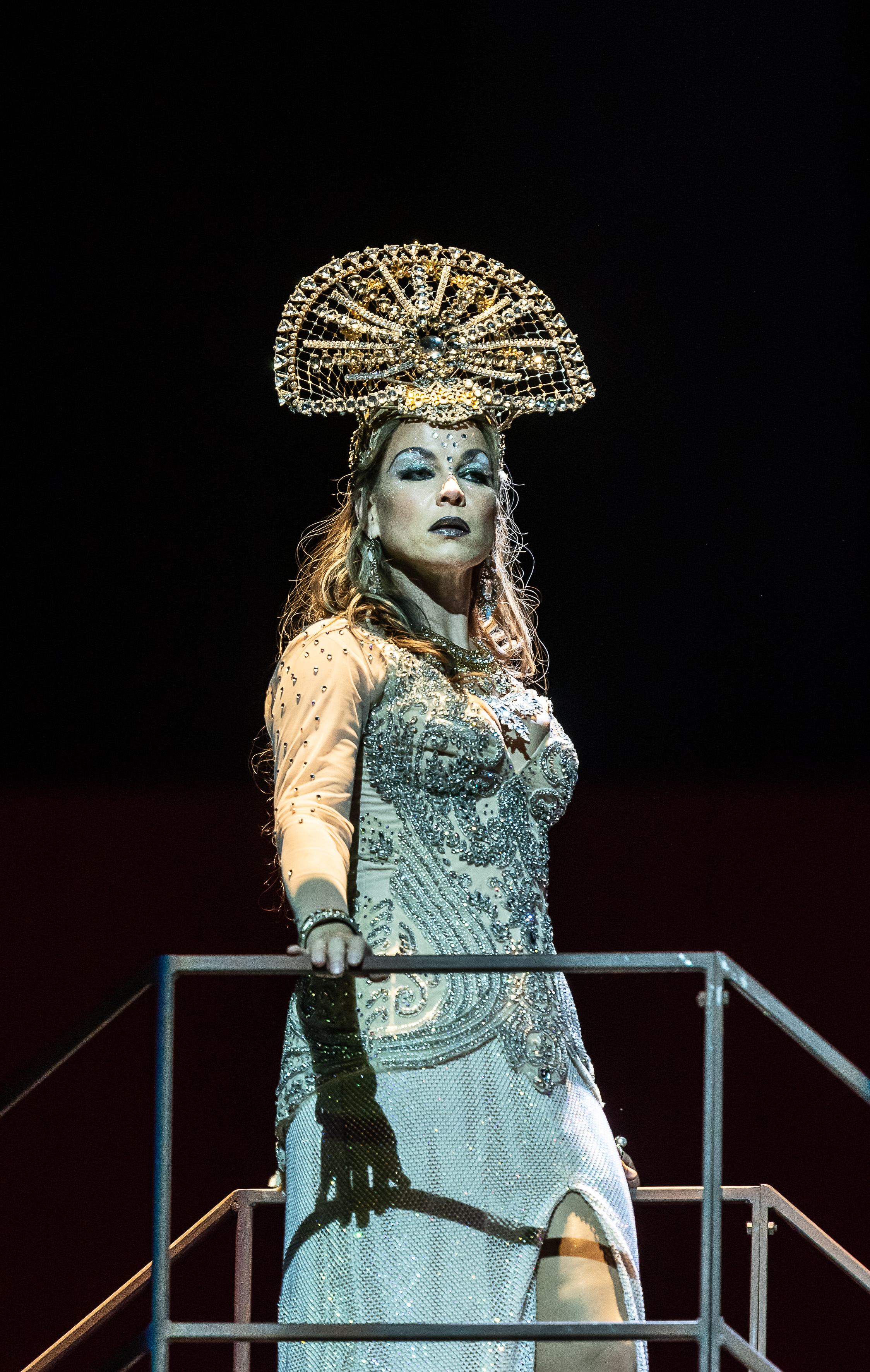 The mezzo-soprano singer Elina Garanča performs as Dalila in The Royal Opera’s production of Samson et Dalila. She stands with one hand on a metal bar and wears a blue beaded dress that splits in the centre to her knee, with a half-moon shaped beaded headdress. Her makeup is vivid with glittery blue eye makeup and black lipstick.  