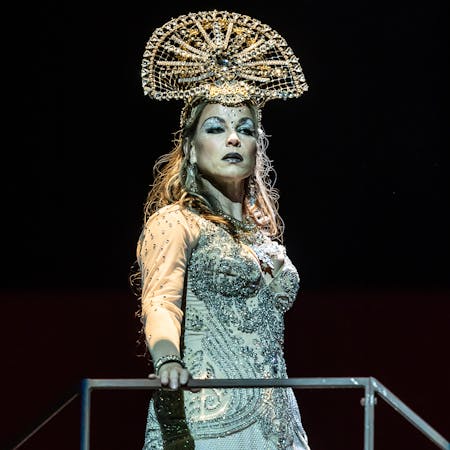 The mezzo-soprano singer Elina Garanča performs as Dalila in The Royal Opera’s production of Samson et Dalila. She stands with one hand on a metal bar and wears a blue beaded dress that splits in the centre to her knee, with a half-moon shaped beaded headdress. Her makeup is vivid with glittery blue eye makeup and black lipstick.