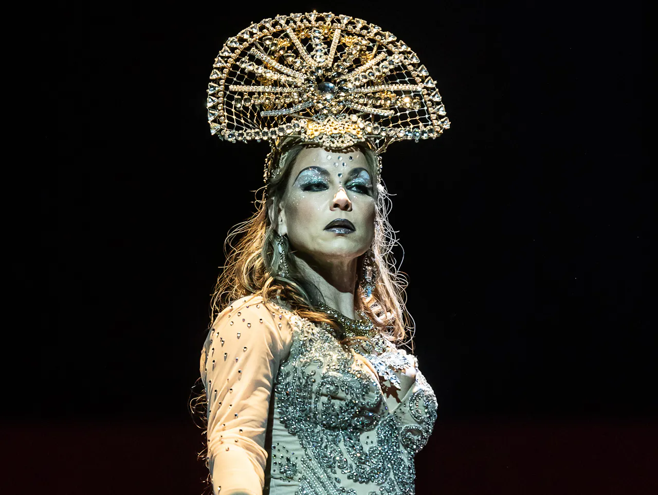 The mezzo-soprano singer Elina Garanča performs as Dalila in The Royal Opera’s production of Samson et Dalila. She stands with one hand on a metal bar and wears a blue beaded dress that splits in the centre to her knee, with a half-moon shaped beaded headdress. Her makeup is vivid withglittery blue eye makeup and black lipstick.